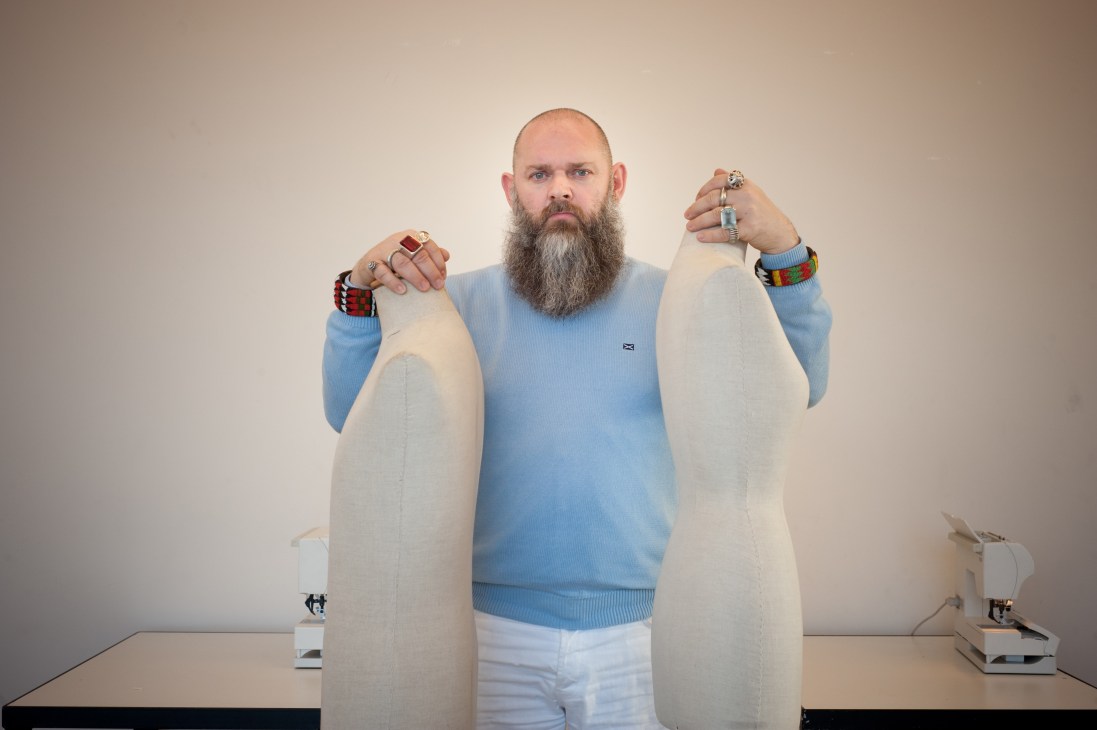 Man with beard wearing blue sweater displaying rings and bracelets while holding cream-colored fabric pieces.