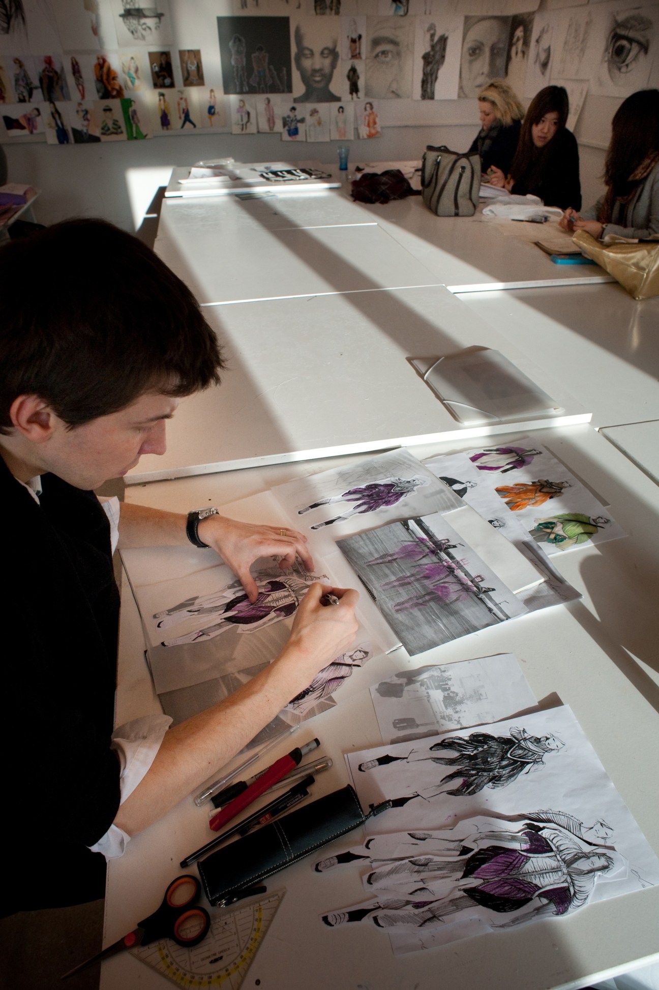 Fashion design students sketch garment designs at a light table in an academy studio.