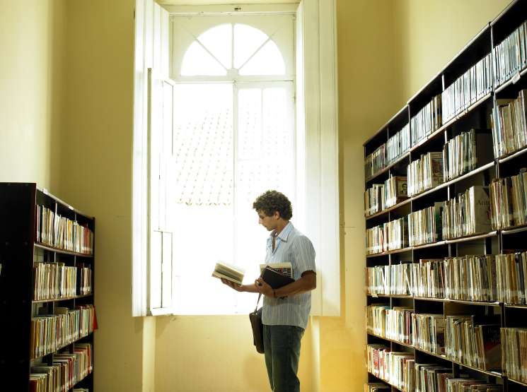 Man reading a book in a bright, yellow-walled library corridor with black bookshelves on both sides.