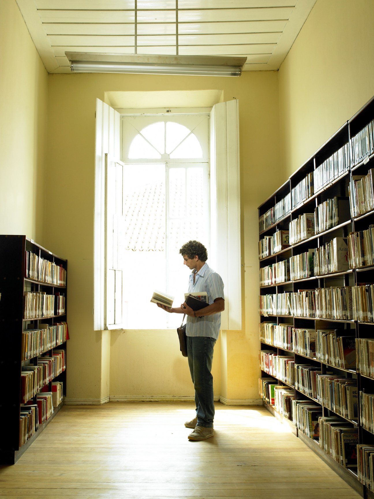 Man reading a book in a bright, yellow-walled library corridor with black bookshelves on both sides.