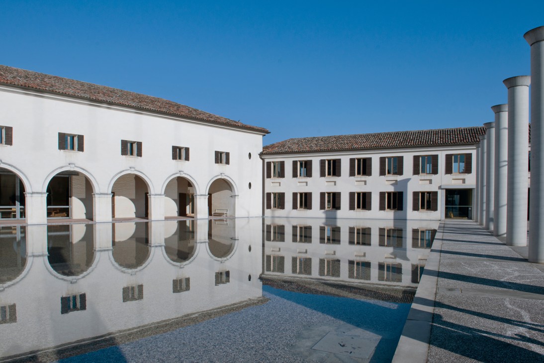 Fabrica headquarters courtyard with white arcaded buildings reflected in water under blue sky.