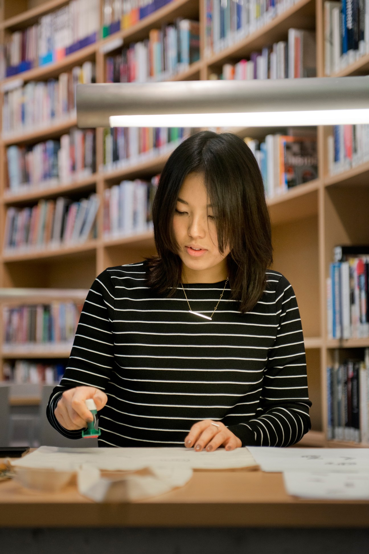 Japanese designer Tomomi Maezawa stamps her name onto a cotton tote bag in front of library shelves.