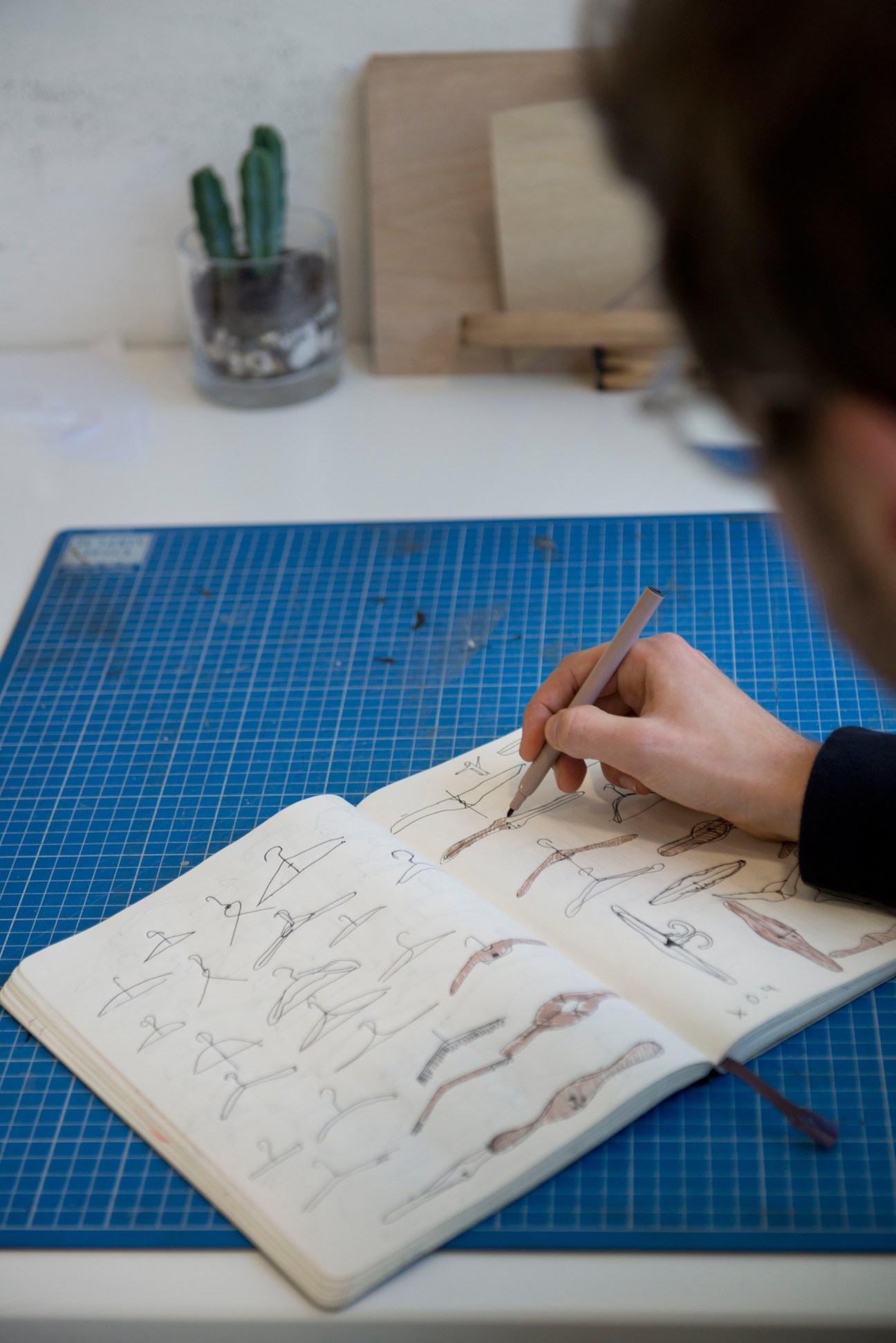 Designer's hand sketching furniture designs in a notebook on a blue cutting mat.