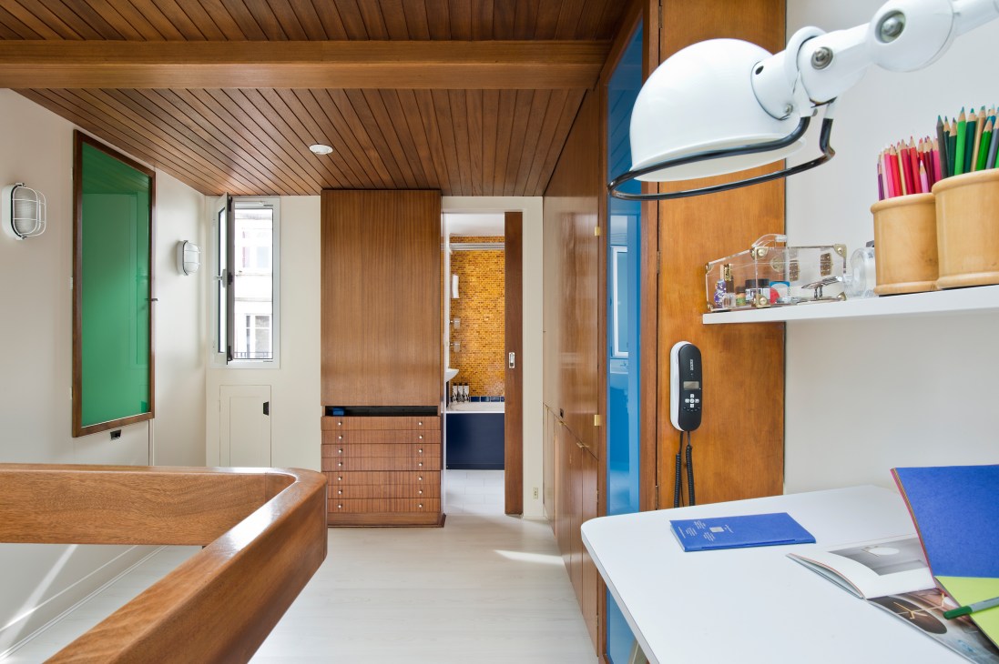 Modern studio apartment interior showing mahogany cabinetry, primary-coloured doors, wooden ceiling, and workspace with shelving and art supplies.