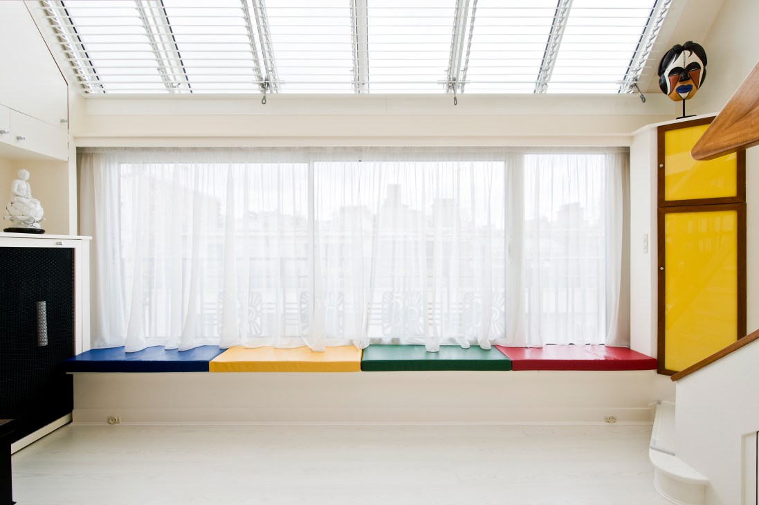 Window seat with primary-coloured storage boxes in blue, yellow, green and red under large windows with white curtains.