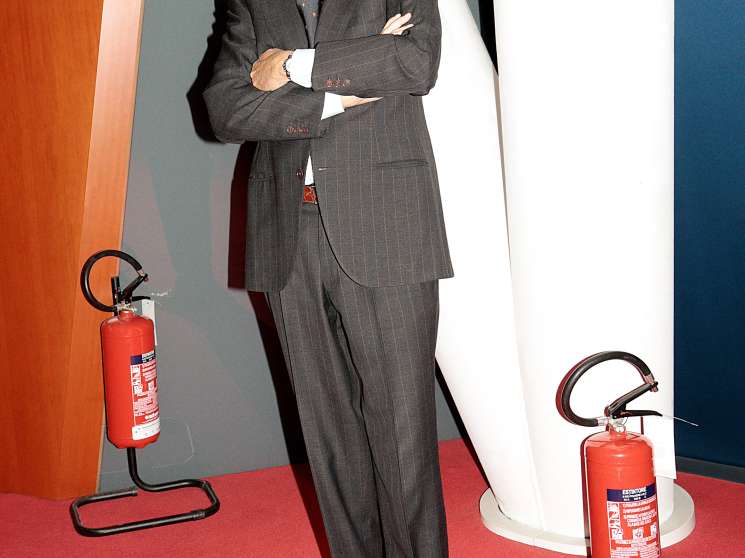 Pirelli CEO Marco Tronchetti Provera stands with arms crossed in a dark pinstriped suit against a geometric background with fire extinguishers.