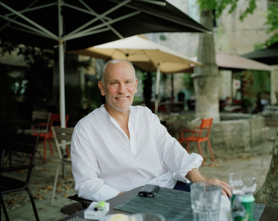 John Malkovich seated at a table on a shaded terrace at Le Fournil restaurant in Bonnieux, Provence.
