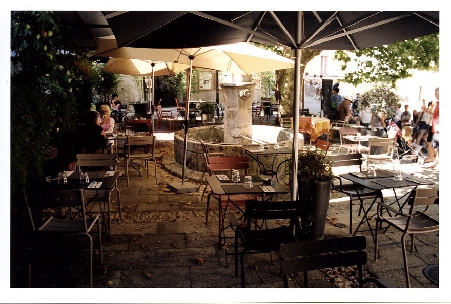 Shaded terrace at Le Fournil restaurant with tables, chairs, umbrellas and diners in Bonnieux.