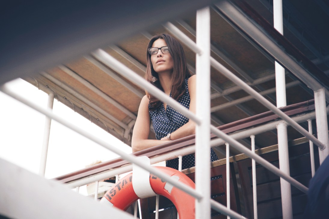 Woman wearing glasses and polka-dot dress on a ferry railing against architectural background.