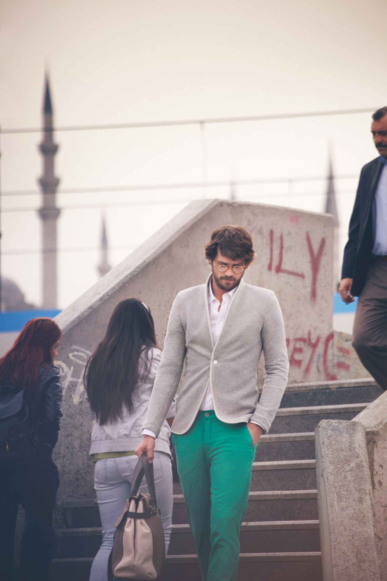 Man wearing Michel Henau glasses, 1205 jacket, Louis Vuitton shirt, Incotex trousers and Yves Saint Laurent bag on waterfront steps with mosque minarets in background.