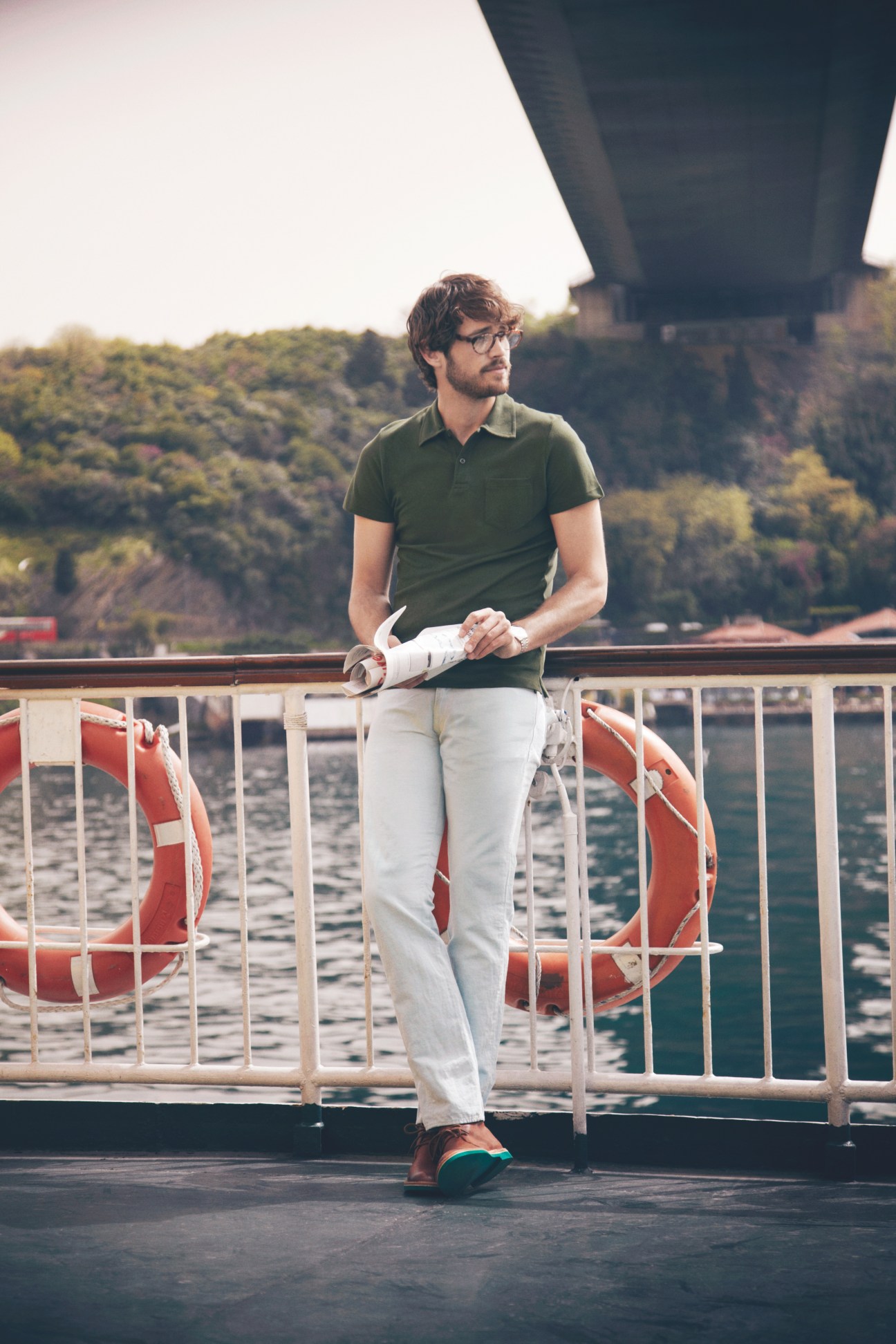 Man in green polo shirt and white jeans leaning on ferry railing with wooded shoreline behind.
