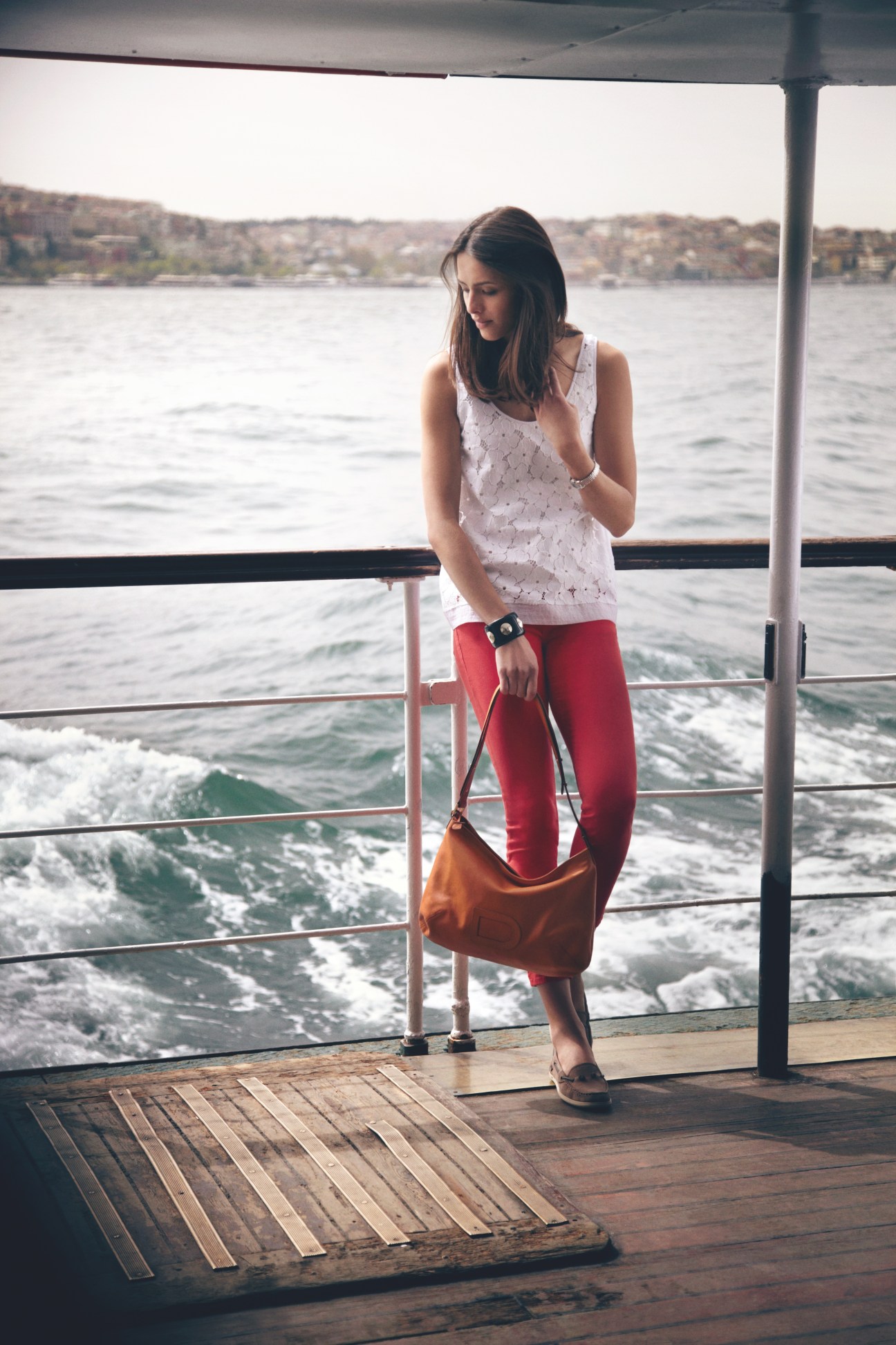 Woman on a ferry wearing a white tank top, red jeans, and brown bag, looking down at the water.