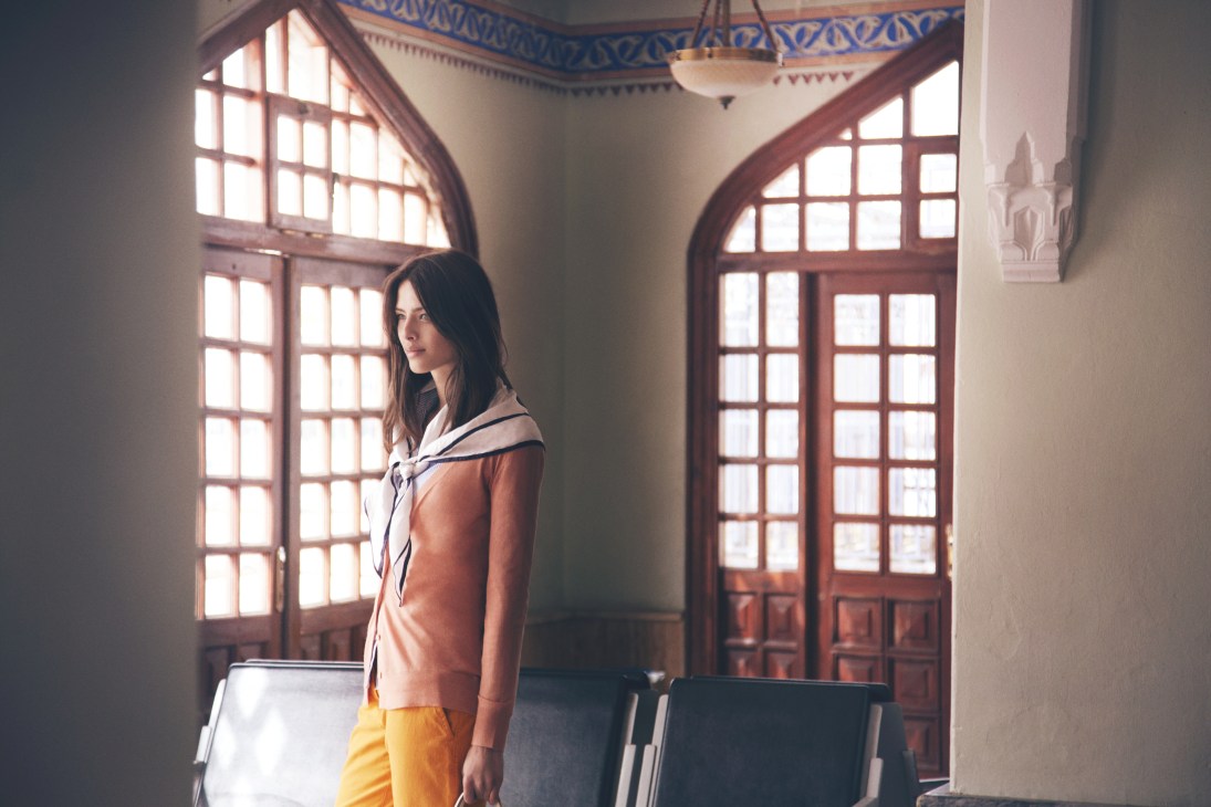 Model wearing Windsor scarf and trousers, Zanone cardigan, J.Crew shirt, and Tod's bag in a sunlit room with arched wooden doors.