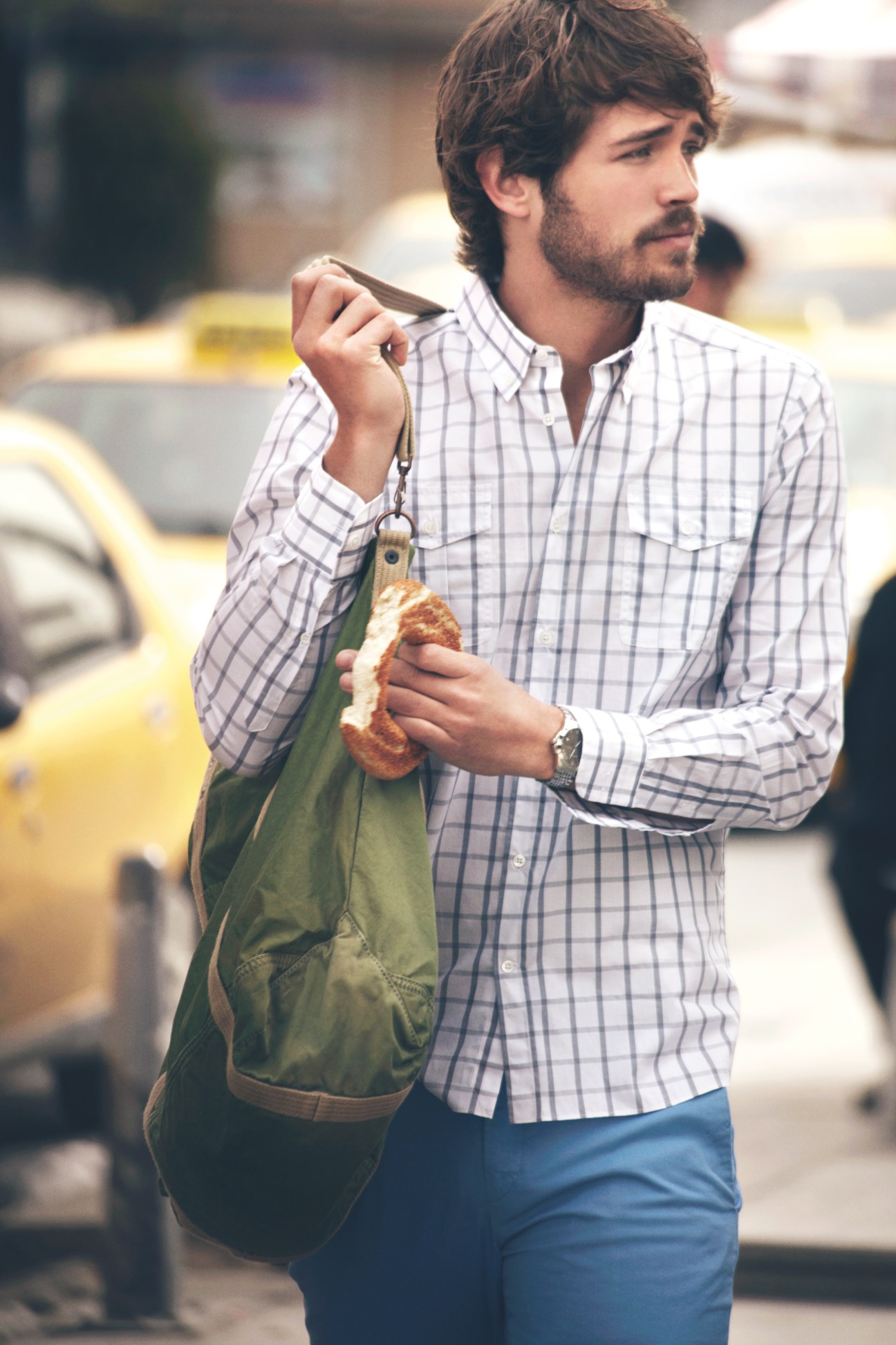 Man in checkered shirt and blue trousers holding green bag and pastry on street.