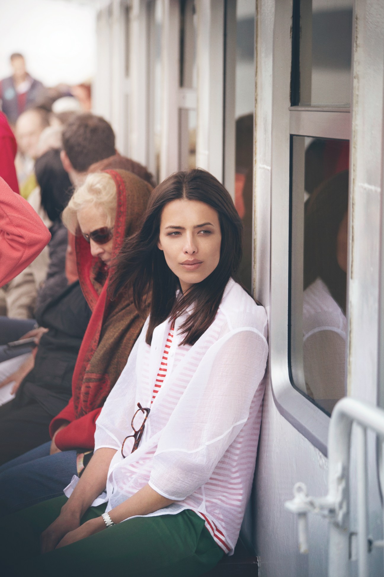 Woman in pink striped shirt and green trousers sits on a ferry with other passengers behind her.