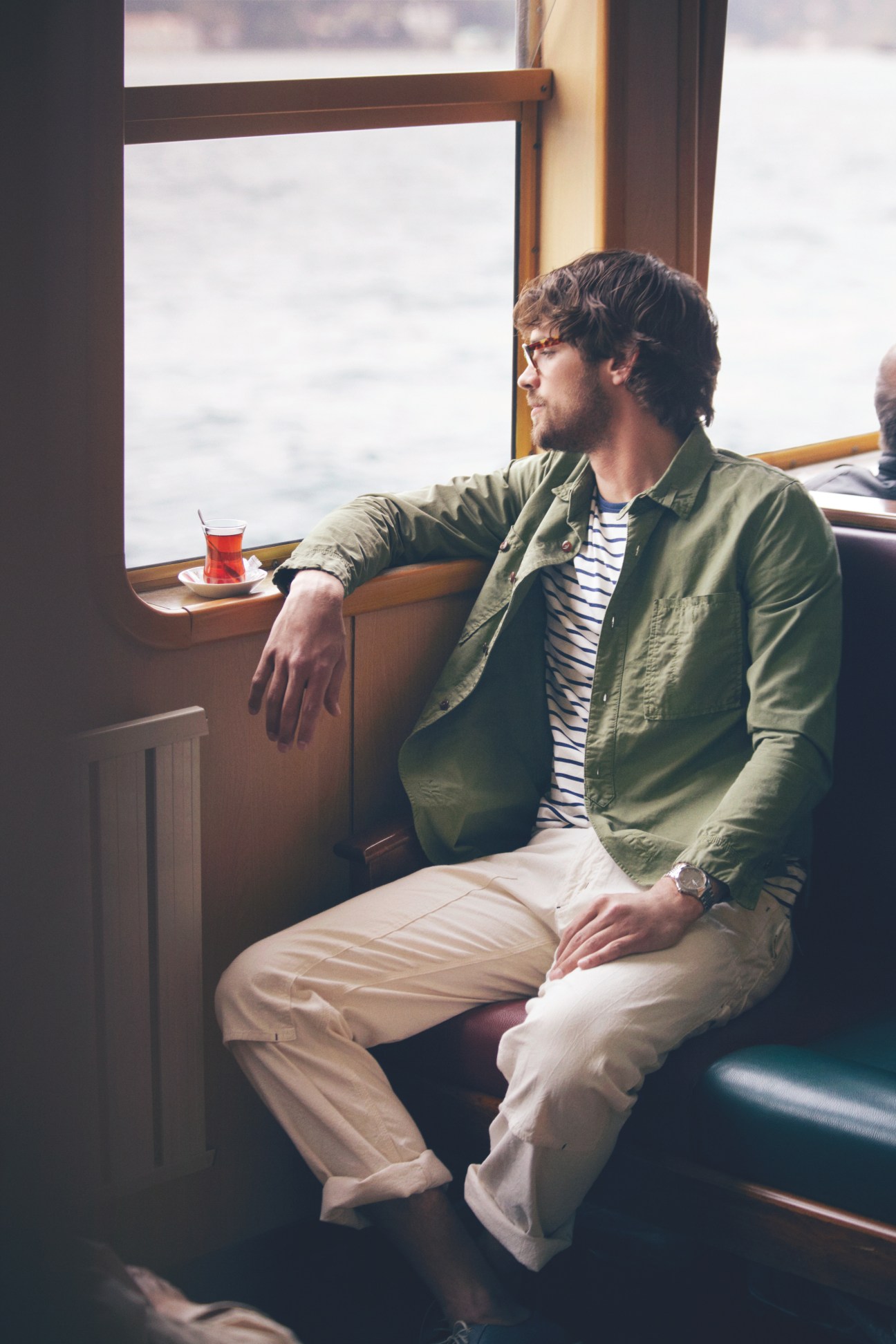 Man wearing Michel Henau glasses and Gucci watch sits by ferry window with tea.