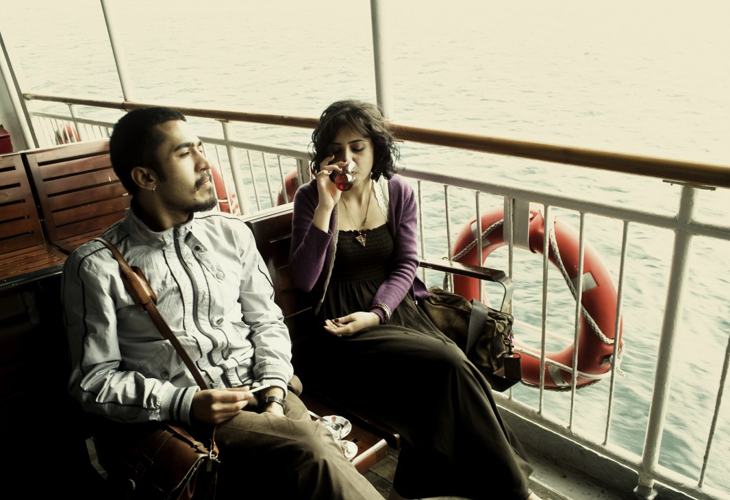 Two passengers sit on an Istanbul ferry deck with tea, overlooking the water.
