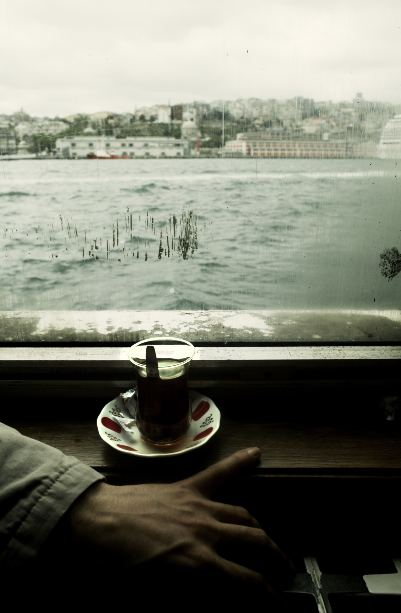 A hand holding a glass of black tea on a saucer by a window overlooking water with buildings and trees.
