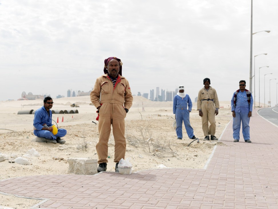 Migrant workers in work clothes stand on a paved area with Doha's skyline visible in the desert background.