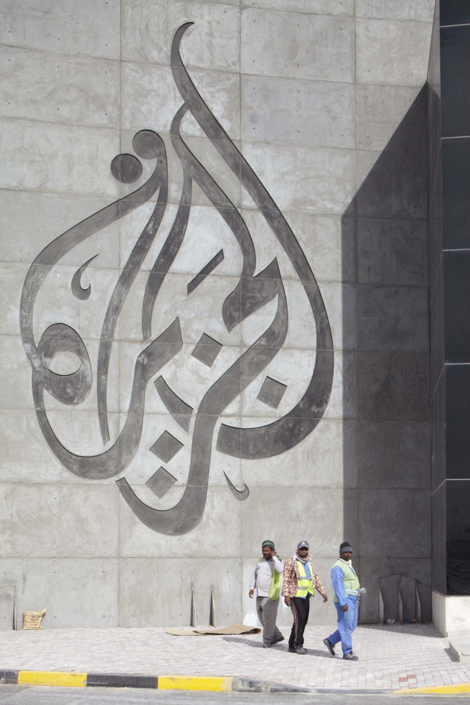 Three migrant workers in safety vests walk past a large Arabic calligraphy mural on an Al Jazeera building wall in Doha.