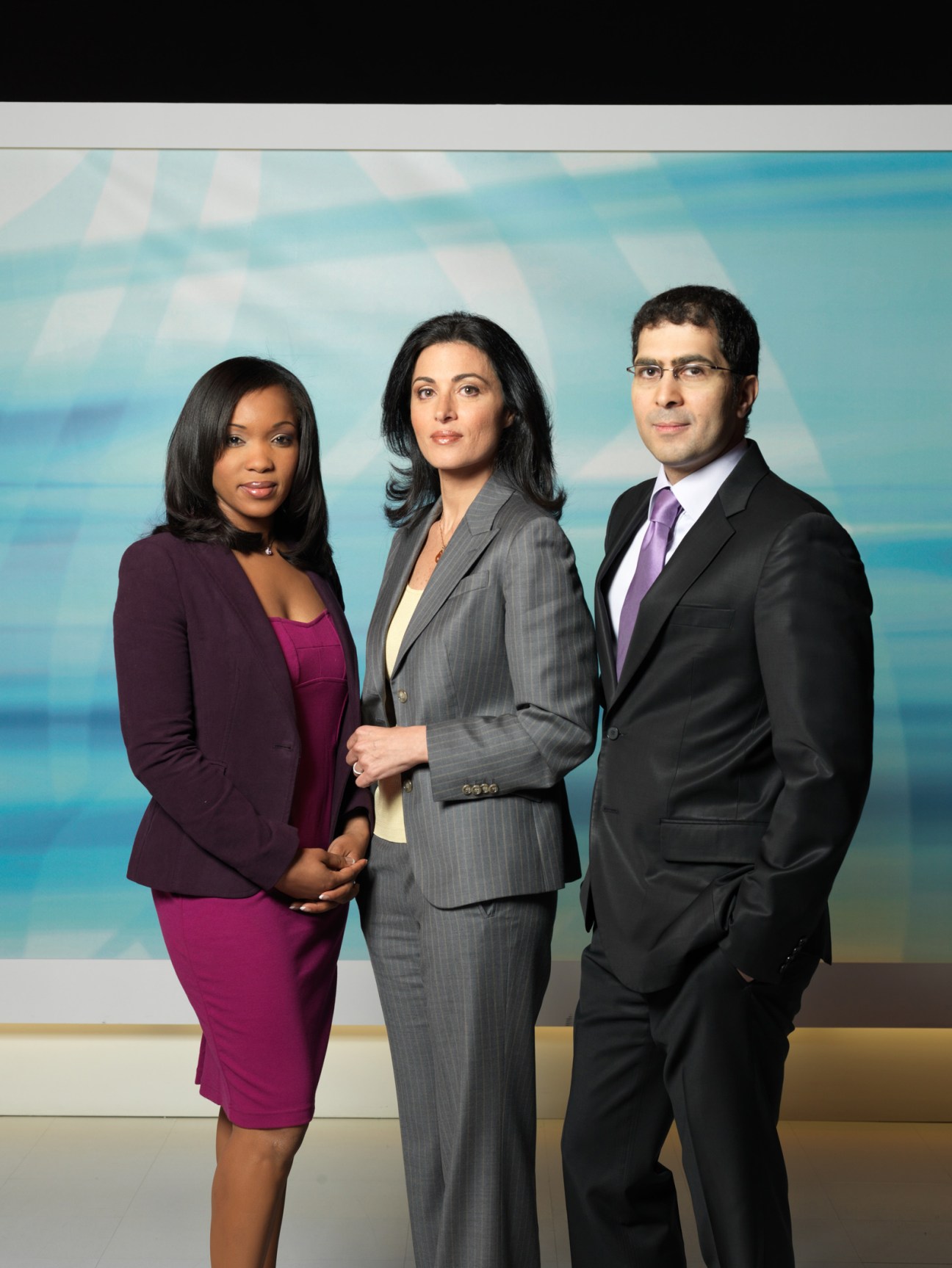 Three Al Jazeera English news anchors pose in professional business attire before a blue gradient studio backdrop.