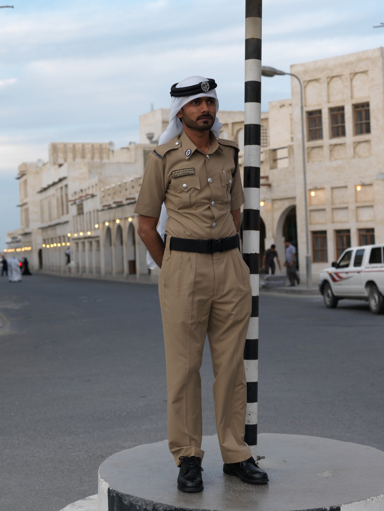 A traffic policeman in traditional Qatari dress stands next to a striped pole on a Doha street.