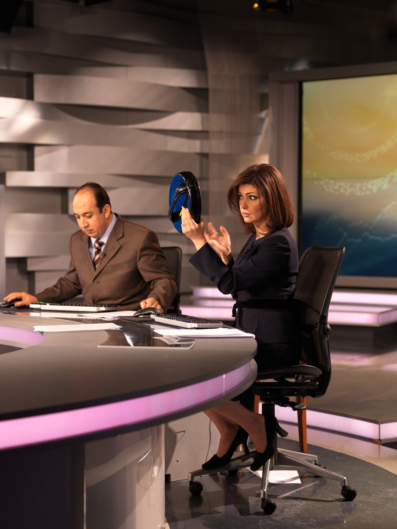 A woman in a dark blazer holds a makeup mirror while sitting at a news desk with a male colleague in a brown suit.