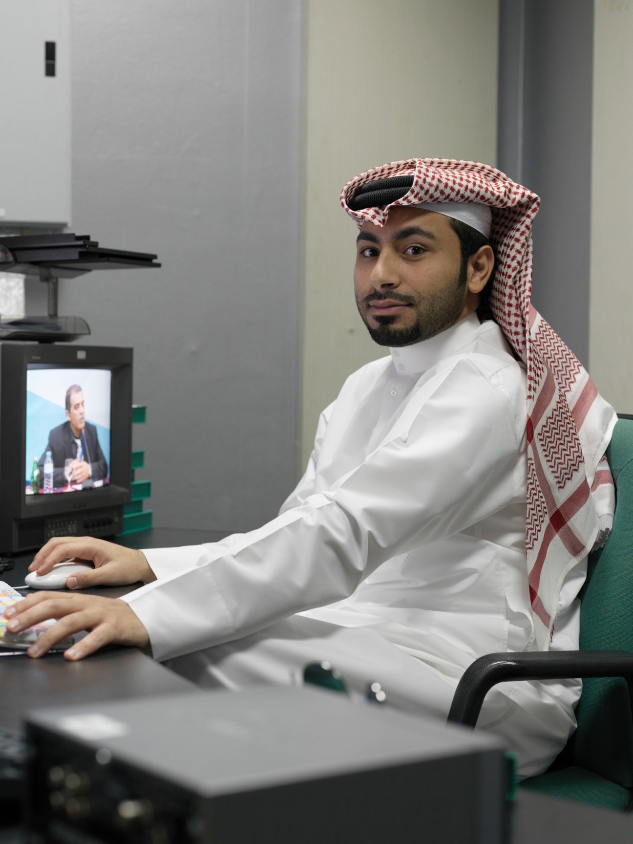 Man in traditional white dishdasha and red-and-white ghutra at desk with computer monitor showing broadcast footage.