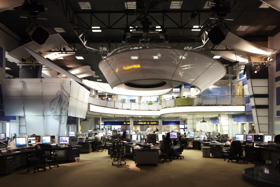 Al Jazeera Arabic newsroom with multiple workstations, computers, and a circular studio set overhead.