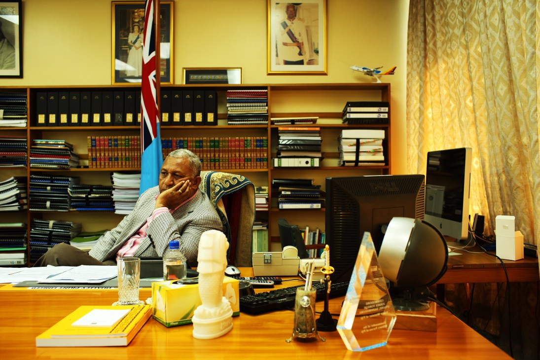 Man in suit sitting at desk in office with bookshelves, Fiji flag, and portraits on yellow wall.