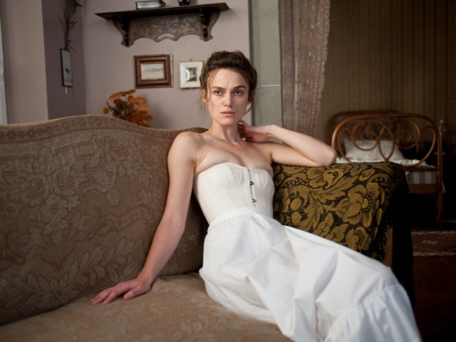 Woman in white strapless dress reclining on a patterned sofa in an ornate interior setting.
