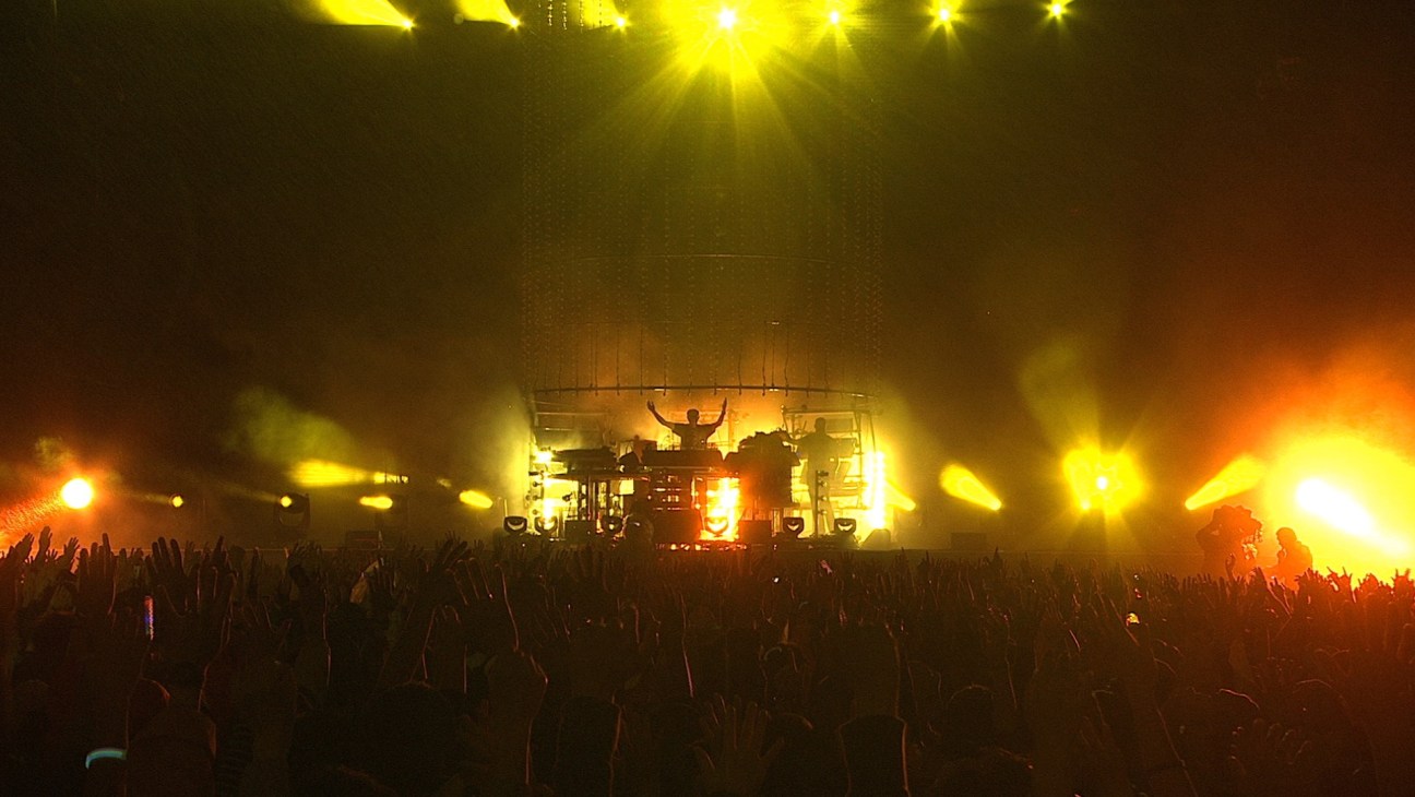 A concert stage with bright yellow and orange lighting illuminating a performer with raised arms above a large crowd.