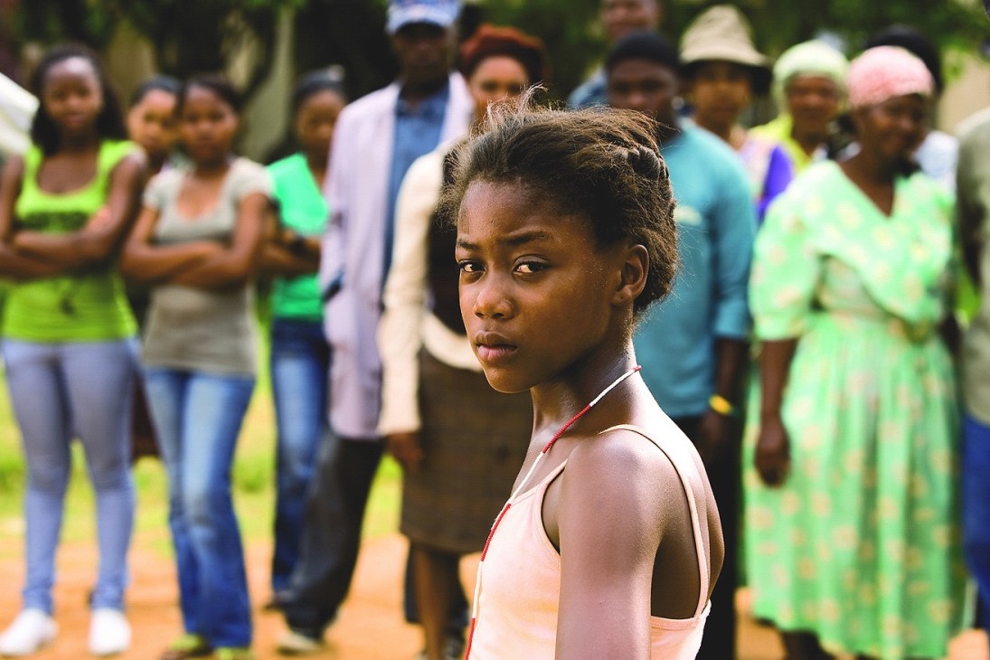 Young girl in pink tank top looks at camera while standing in front of a blurred crowd of onlookers outdoors.