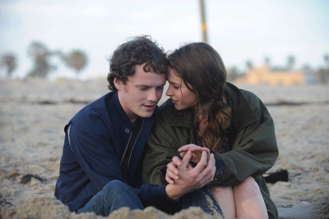 A young couple sits together on a beach, gazing at each other intimately while sitting in the sand.