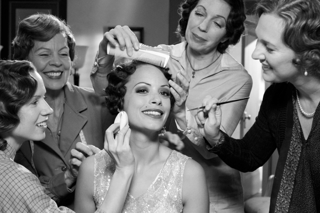 Black and white scene from 1920s Hollywood film showing a woman receiving makeup and hair styling from multiple attendants.