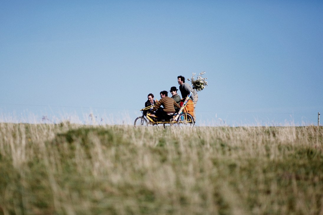 Five people on a yellow cargo bike riding through a grassy coastal landscape under blue sky.