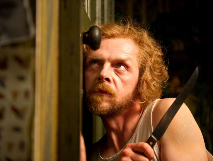 A man with reddish hair and beard wearing headphones around his neck, holding a knife, with an anxious expression.