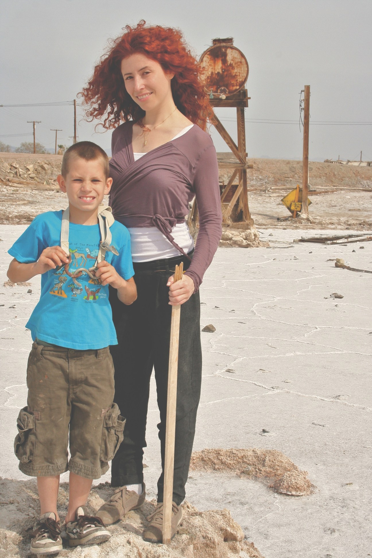 Woman with red curly hair and young boy standing in desert landscape with abandoned industrial equipment behind them.