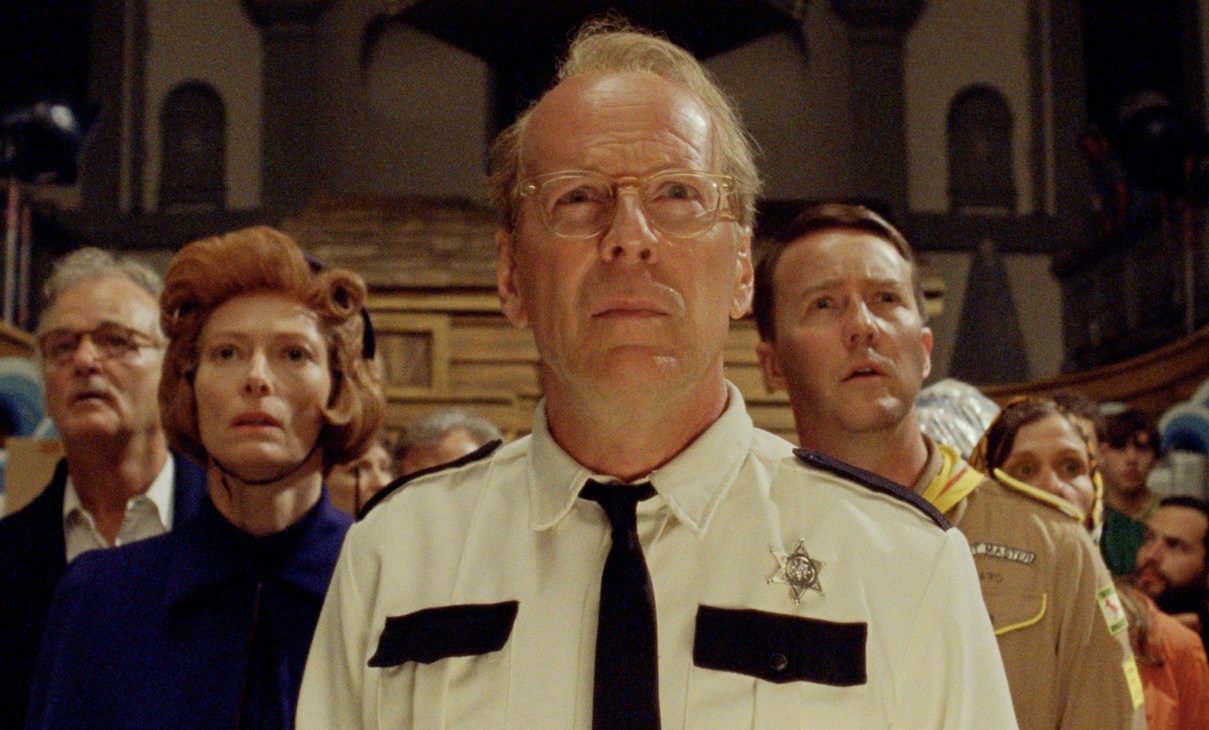 A man in glasses and a light-colored uniform with black trim stands among other people in an ornate interior setting.