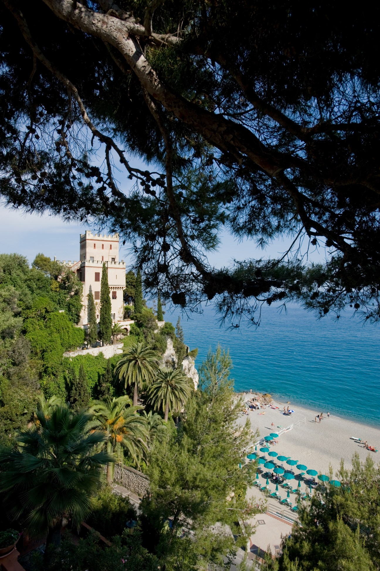 A stone tower overlooks a sandy beach with turquoise water and palm trees in Finale Ligure, Italy.