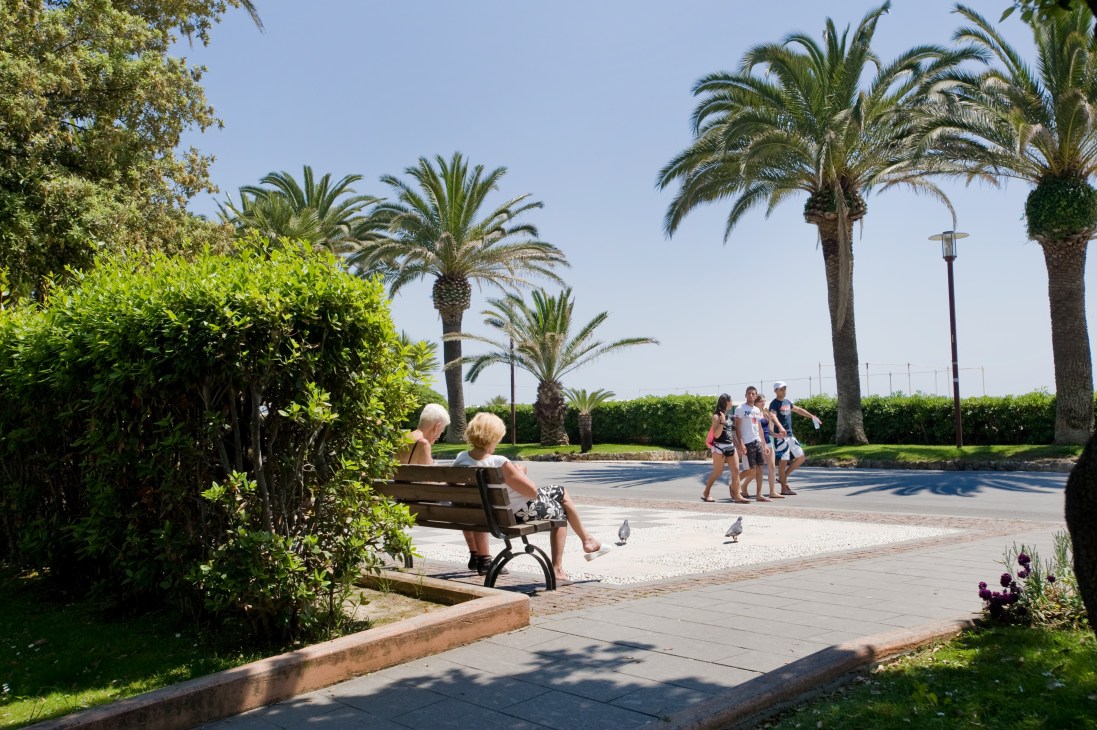 Palm-lined promenade in Finale Ligure with beachgoers and sandy shore.