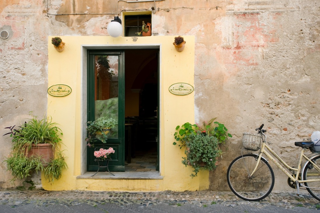 Yellow-painted storefront with green door, potted flowers, and bicycle parked outside in Finale Ligure.