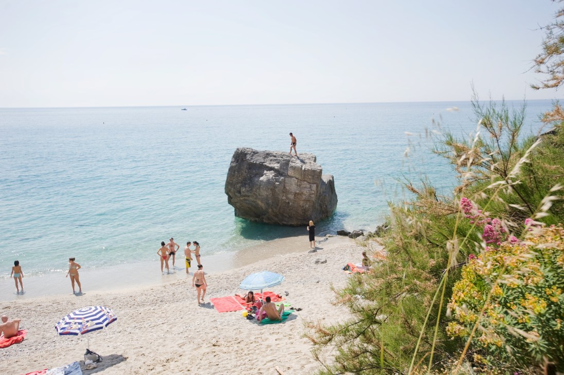 Beach scene at Finale Ligure with swimmers, sunbathers under umbrellas, and a large rock formation in the sea.