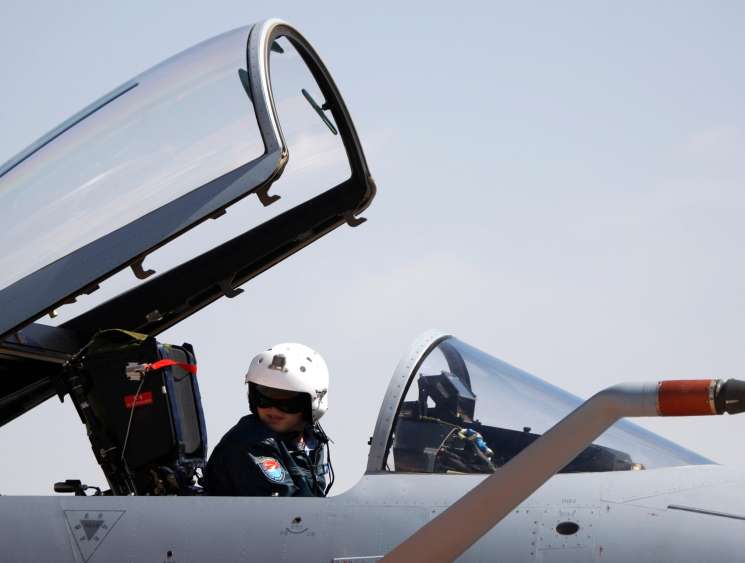 Fighter jet pilot in white helmet sitting in open cockpit of military aircraft with canopy raised against clear sky.