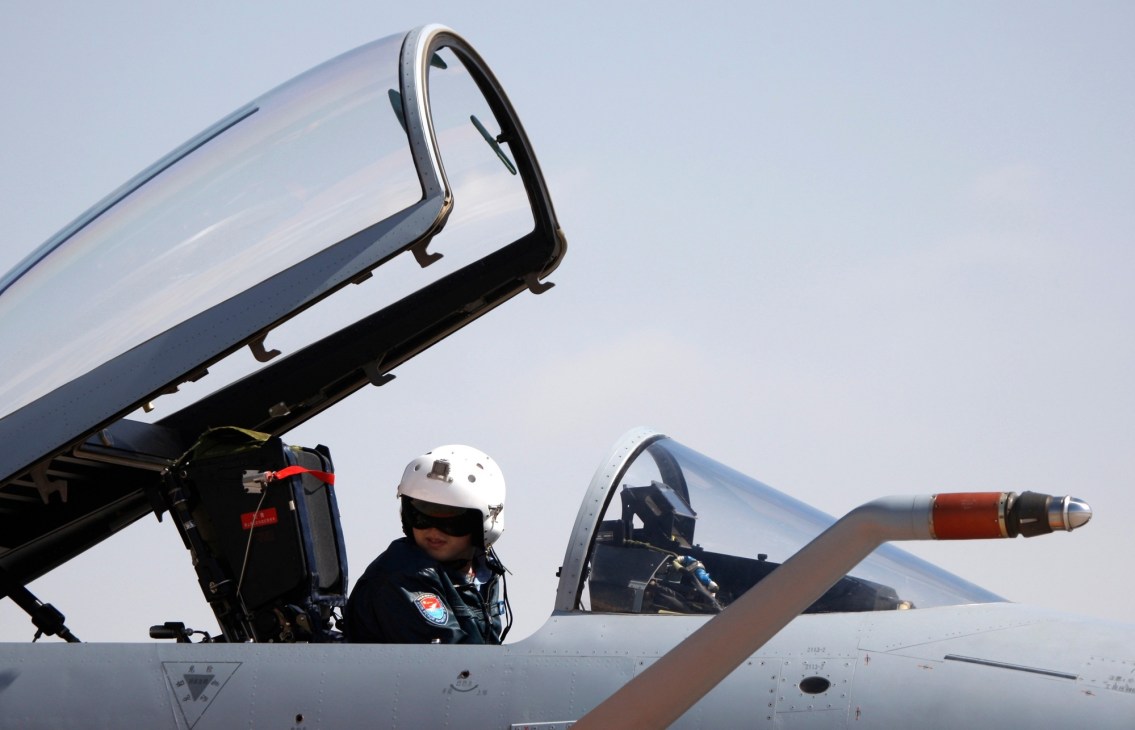 Fighter jet pilot in white helmet sitting in open cockpit of military aircraft with canopy raised against clear sky.