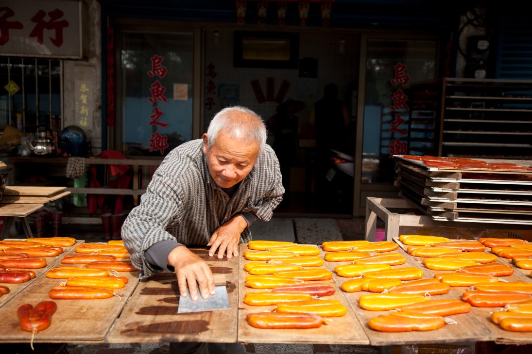 An elderly man arranges orange mullet roe on wooden drying racks outside a shop with Chinese signage.
