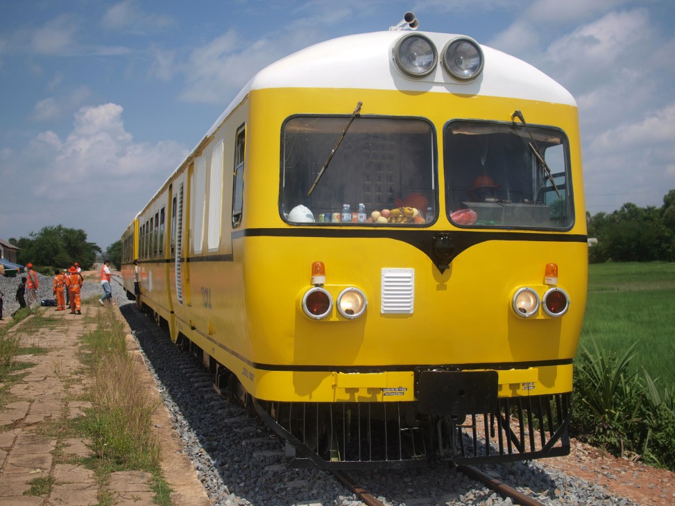 A bright yellow vintage train with white roof and dual headlights on railway tracks surrounded by green fields.