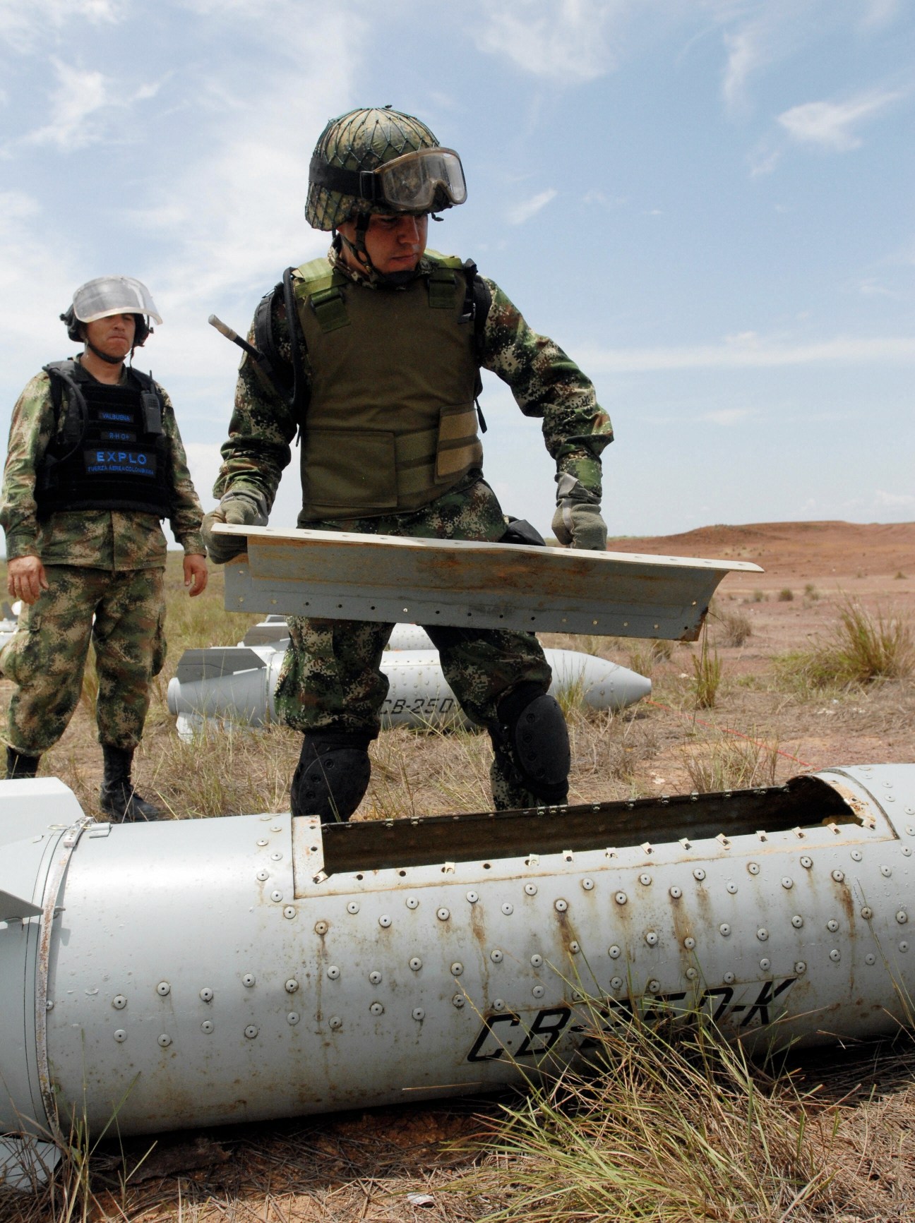 Two soldiers in camouflage uniforms examining a metal cylindrical object in an arid landscape.