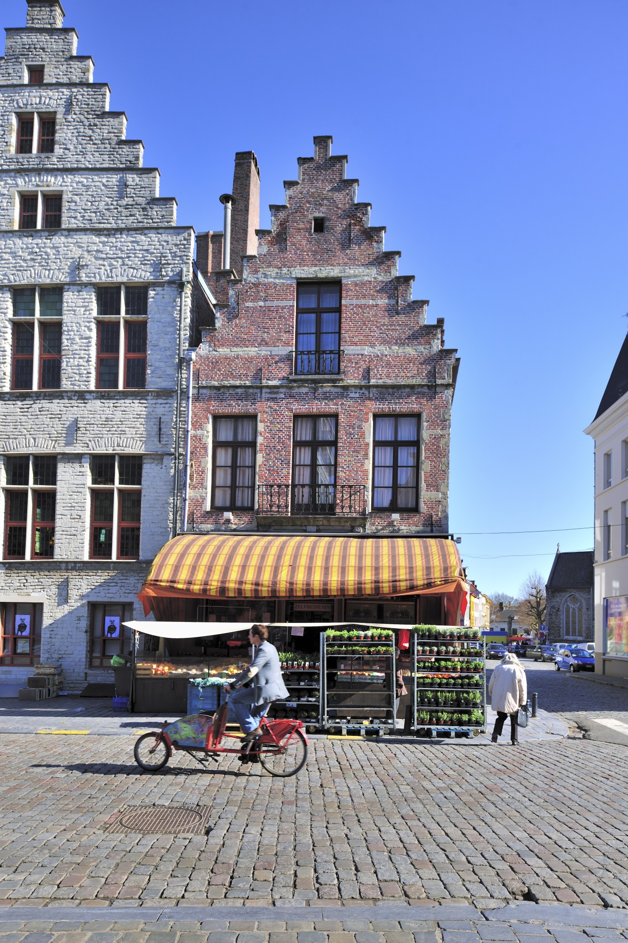 Cyclist riding past Fruitco fruit and vegetable shop with striped awning on a sunny Ghent street.