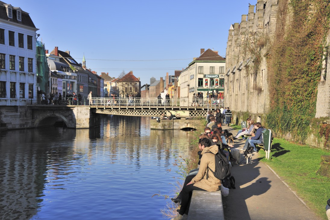 People relaxing by the Leie river in Ghent with historic buildings and a bridge reflected in the water.